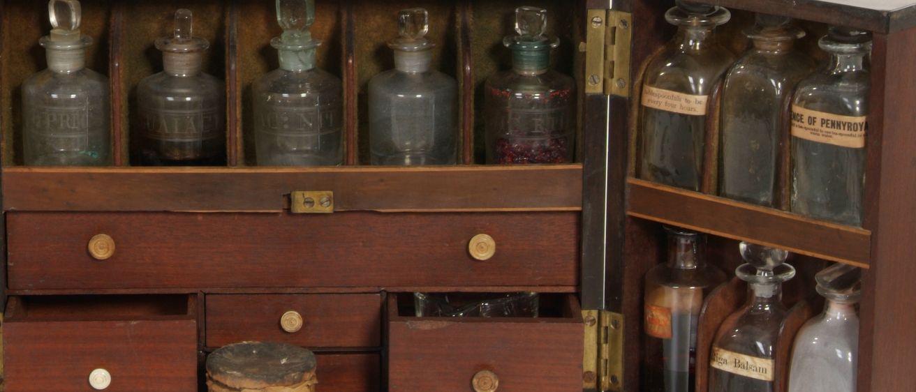 Ein Vintage-Medizinkasten aus Holz mit Messingbeschlägen, der verschiedene Glasflaschen und Gläser mit Etiketten enthält, in mehreren Schubladen und Fächern angeordnet, auf einer weißen Oberfläche platziert.