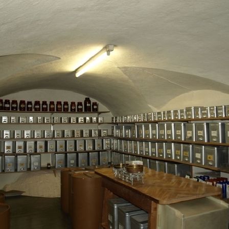 Ein Raum mit hoher Decke und vielen Regalen mit verschiedenen Gegenständen darauf. Die Regale enthalten zahlreiche Behälter, darunter Metallboxen und Glasflaschen. Eine Lampe ist an der Decke montiert.