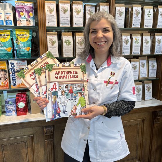 Eine Frau in einem weißen Laborkittel lächelt, während sie ein Buch über Apotheken in den Händen hält.