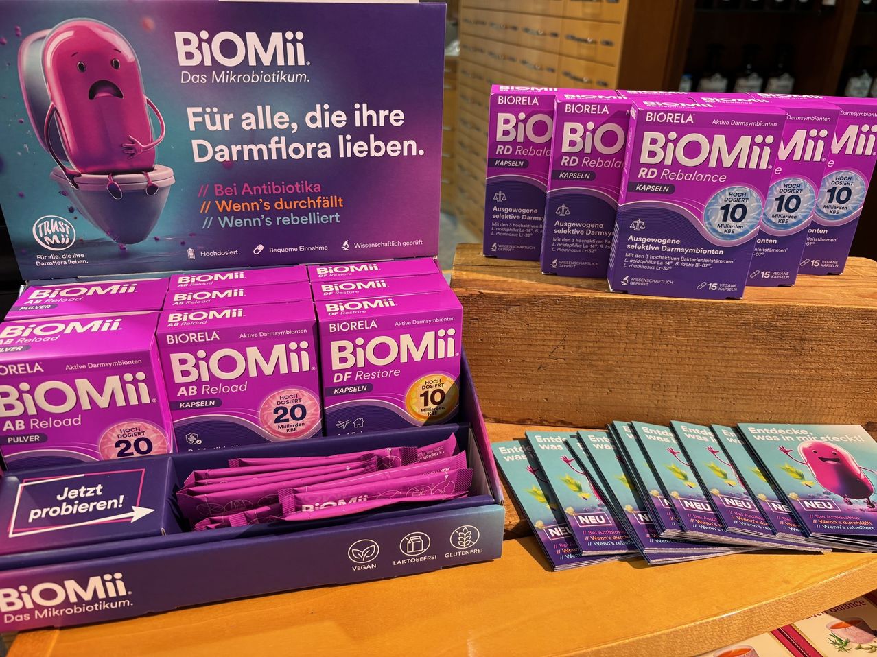 Eine Anzeige von BioMii Probiotika-Ergänzungsmitteln in verschiedenen Packungen auf einem Holzregal. Die Hauptpackung zeigt eine Zeichentrickfigur und Text über das Produkt.