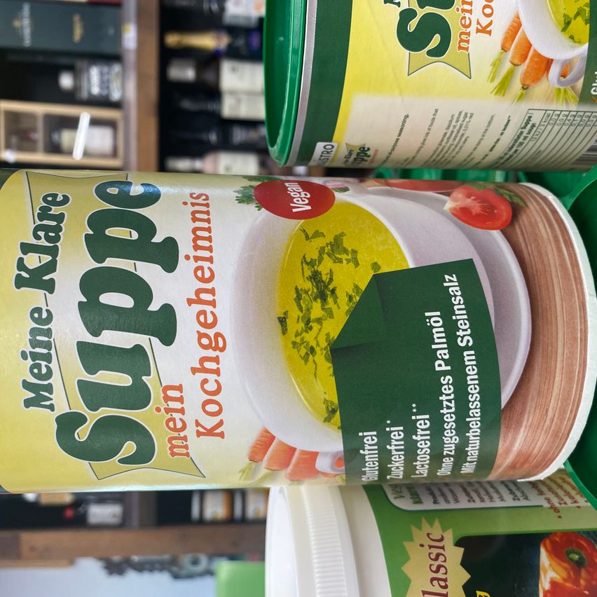 Eine Dose Suppe mit der Aufschrift 'Suppe mein Kochgeheimnis' und einem grünen Etikett, das 'glutenfrei' und 'lactosefrei' anzeigt. Es zeigt eine Tomaten- und Karottensuppe mit Kräutern.
