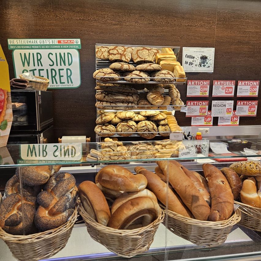 A bakery display case filled with various types of bread and pastries, including pretzels, rolls, and cookies. The sign reads 'Wir sind steirer', indicating the bakery's regional identity.