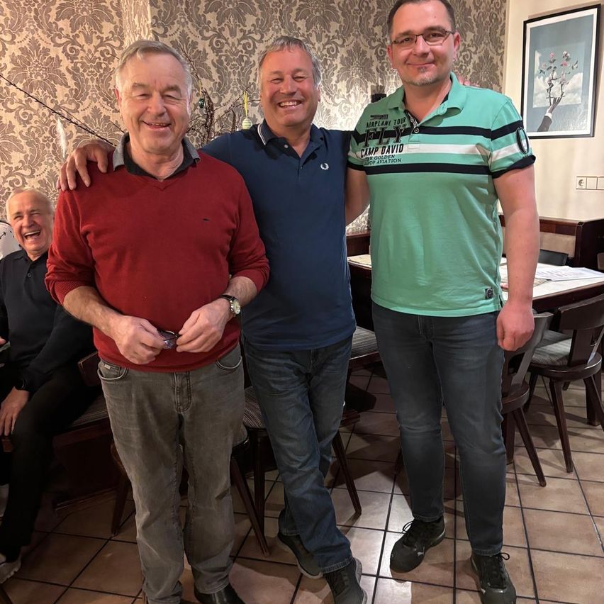 Drei Männer stehen in einem Restaurant und lächeln für ein Foto. Einer trägt einen roten Pullover, der andere ein blaues Poloshirt und der dritte ein grünes Poloshirt mit Camp David. Ein vierter Mann sitzt im Hintergrund.