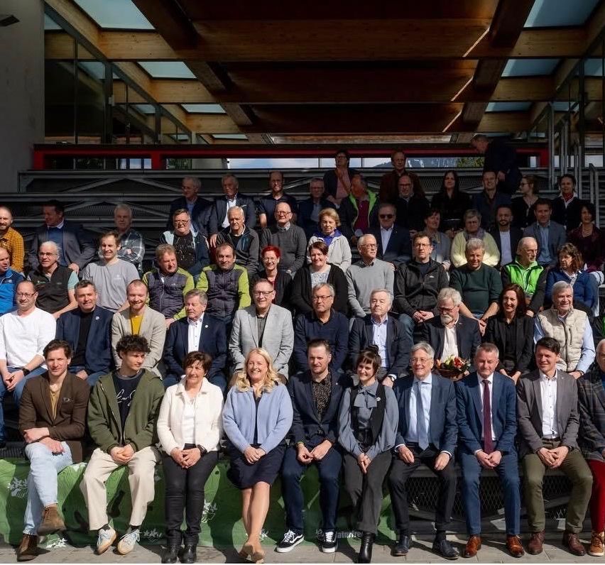 Eine große Gruppe von Menschen in formeller Kleidung sitzt und steht auf Tribünen und posiert für ein Gruppenfoto unter einer Holzstruktur.