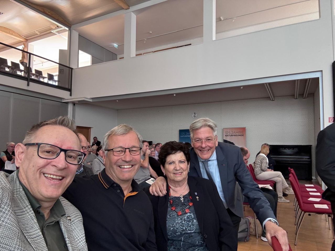 Vier Personen lächeln und posieren für ein Foto in einem Saal mit Holzboden und weißen Wänden. Zwei Männer und zwei Frauen sind formell gekleidet.