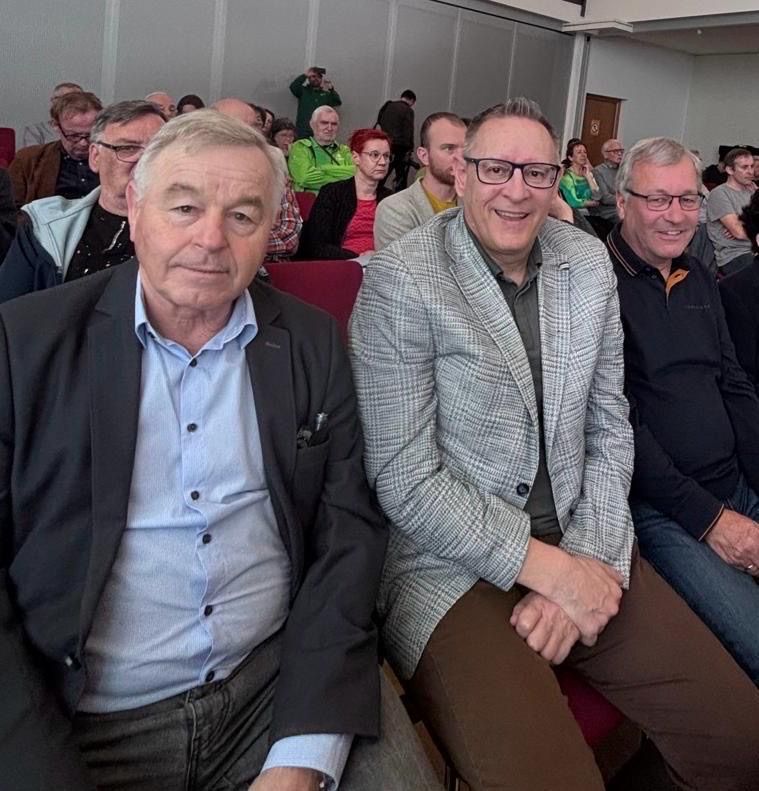 Eine Gruppe von Menschen sitzt in einem Auditorium. Drei Männer lächeln vorne, während andere beobachten. Einige tragen Brillen, andere haben Kameras.