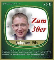Ein grünes Etikett zeigt einen lächelnden Mann mit Brille. Über seinem Gesicht steht 'Zum 30er', darunter 'Hubertus Pils'. Der Hintergrund hat ein hellgrünes und dunkelgrünes Farbschema.