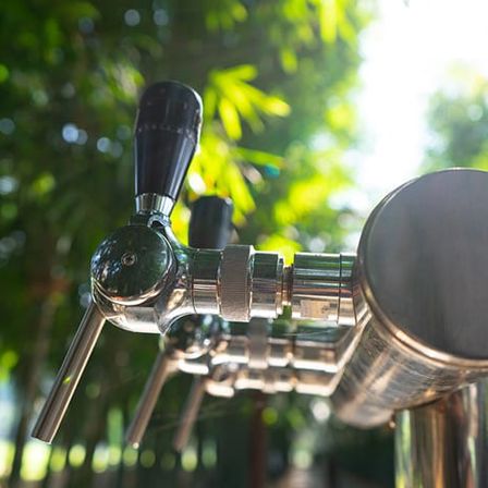 Ein Nahaufnahme eines Edelstahl-Bierhahns mit schwarzem Griff, vor einem Hintergrund aus grünen Bäumen und Sonnenlicht.