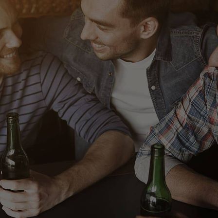 Drei Männer sitzen zusammen, zwei halten grüne Flaschen, lächeln und schauen nach vorne, einer in einer Jeansjacke und ein anderer in einem karierten Hemd.