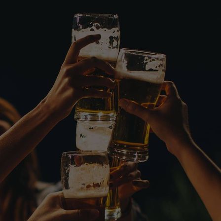 Mehrere Hände, die Gläser mit Bier halten, sind zum Anstoßen gemeinsam erhoben. Die Gläser sind mit schaumigen Bier gefüllt, und die Hände sind verschwommen. Der Hintergrund ist dunkel.