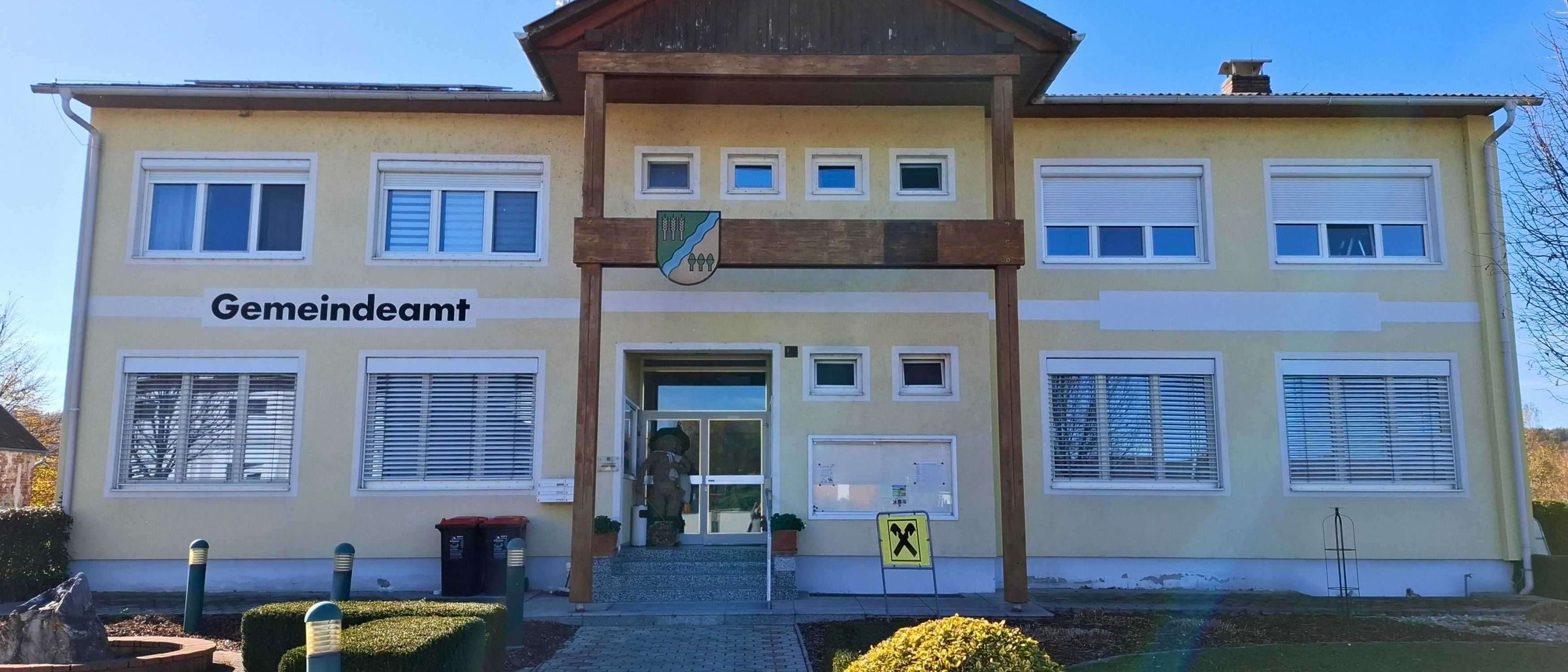 Die Vorderseite eines gelben Gebäudes mit der Aufschrift 'Gemeindeamt'. Es hat eine Holzveranda, Glastüren und ein gelbes Schild mit einem 'X'. Vor dem Gebäude befinden sich Mülleimer, Topfpflanzen und Büsche.