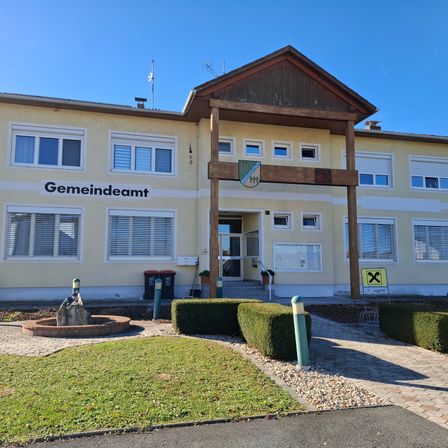 Das Bild zeigt ein Gebäude mit einem braunen Dach und weißen Wänden. Es trägt die Aufschrift Gemeindeamt. Es gibt mehrere Fenster am Gebäude und einige Mülleimer davor. Ein Brunnen steht vor dem Gebäude.
