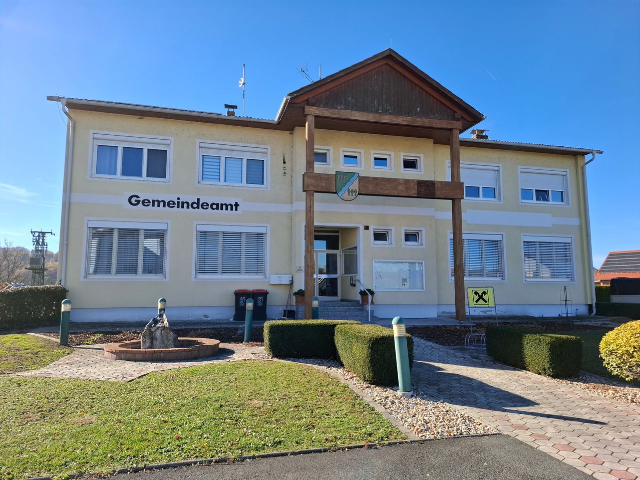 Das Bild zeigt ein Gebäude mit einem braunen Dach und weißen Wänden. Es trägt die Aufschrift Gemeindeamt. Es gibt mehrere Fenster am Gebäude und einige Mülleimer davor. Ein Brunnen steht vor dem Gebäude.