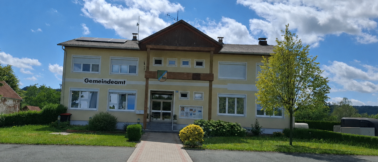 Ein Gebäude mit 'Gemeindeamt' auf der Vorderseite hat einen gepflasterten Weg, der zum Eingang führt. Es hat Solarmodule auf dem Dach.