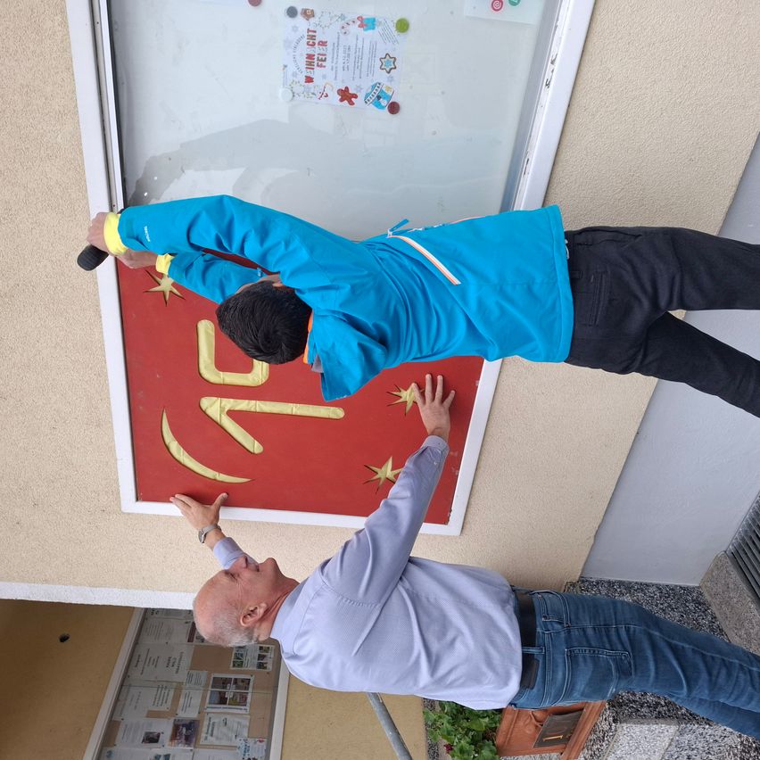 Zwei Männer hängen ein rotes Banner mit chinesischen Schriftzeichen an einer Wand auf, einer hält einen Pinsel.