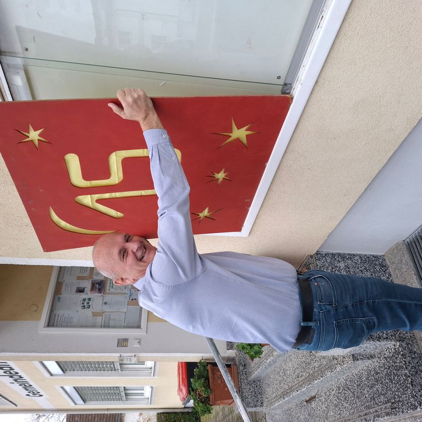 Ein Mann lächelt, während er ein rotes Schild mit einem gelben Sternmuster hochhält, möglicherweise an einem Gebäude befestigt.