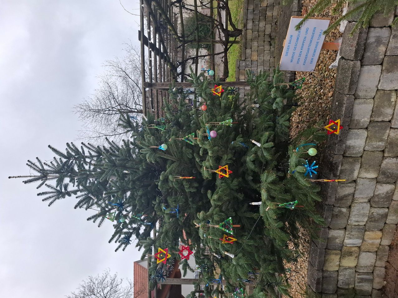 Ein großer geschmückter Weihnachtsbaum steht vor einer Steinmauer, verziert mit bunten Ornamenten und dreieckigen Dekorationen. Ein Schild ist neben dem Baum platziert, mit einem Holzzaun dahinter. Der Hintergrund zeigt einen Grasbereich und einen bewölkten Himmel.