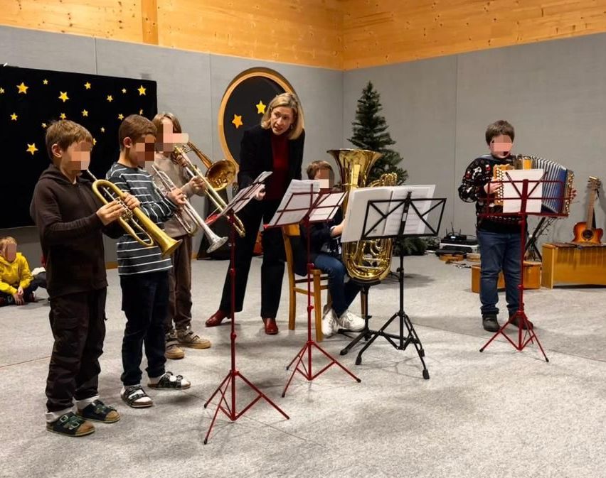 Eine Gruppe von Kindern spielt verschiedene Blechblasinstrumente, während eine Frau, möglicherweise ihre Lehrerin, mit Notenblättern in der Nähe steht. Der Raum ist mit einem Weihnachtsbaum und einem sternförmigen Hintergrund dekoriert.