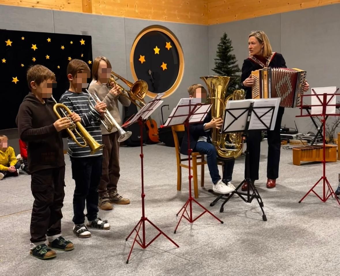 Eine Gruppe von Kindern und einem Erwachsenen übt Musik. Sie stehen mit ihren Trompeten, Tuba und Akkordeon. Dahinter steht ein Weihnachtsbaum und eine Kulisse mit Sternen.