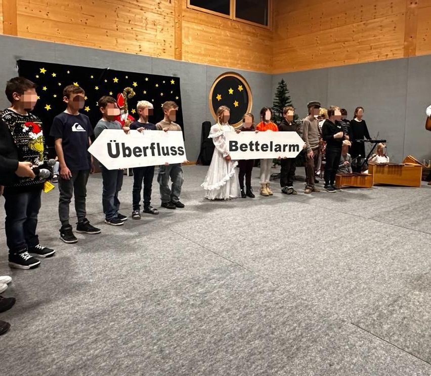 Mehrere Kinder halten Schilder mit den deutschen Wörtern 'Überfluss' und 'Bettelarm'. Sie stehen in einem Raum mit Holzwänden und einem Weihnachtsbaum.