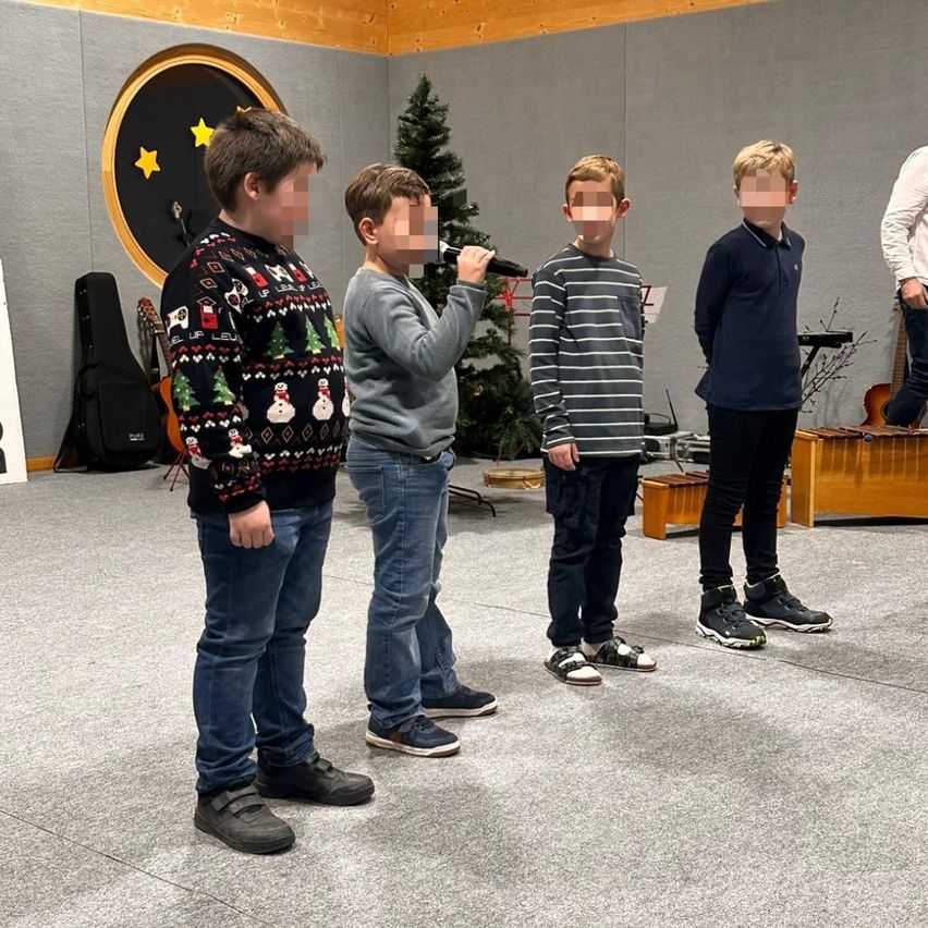 Vier Jungen stehen in einem Raum, einer hält ein Mikrofon. Hinter ihnen sind ein Weihnachtsbaum und Musikinstrumente zu sehen. Sie scheinen sich auf eine Aufführung vorzubereiten.