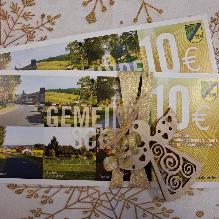 Ein Stapel gelber und goldener Gutscheine mit Text und Bildern, einschließlich '10€', 'GEMEINDE SCH' und einem hölzernen Engel mit Wirbeln. Der Hintergrund zeigt eine ländliche Szene mit einer Straße, Häusern und Bäumen.