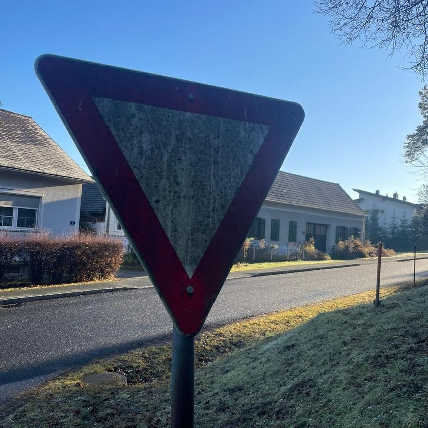 Ein rotes und weißes dreieckiges Straßenschild steht am Straßenrand. Dahinter befindet sich ein Haus mit einem braunen Dach. Der Himmel ist klar und blau.