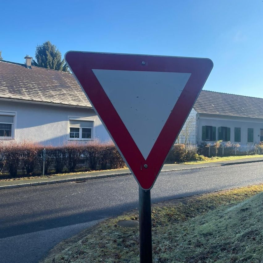 Ein rotes und weißes dreieckiges Verkehrsschild steht am Straßenrand in der Nähe eines Hauses mit einem braunen Dach.