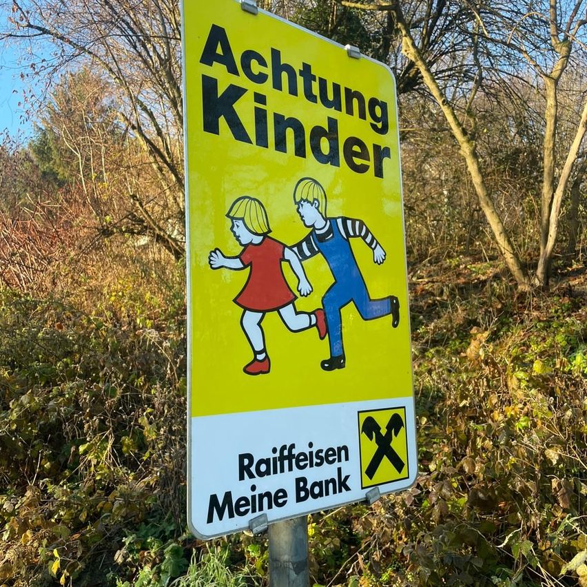 Ein gelbes Schild mit schwarzen Buchstaben warnt vor Kindern. Es zeigt zwei rennende Kinder mit einem durchgestrichenen Symbol am unteren Rand. Das Schild ist auf einem Pfosten in einer natürlichen Umgebung befestigt.