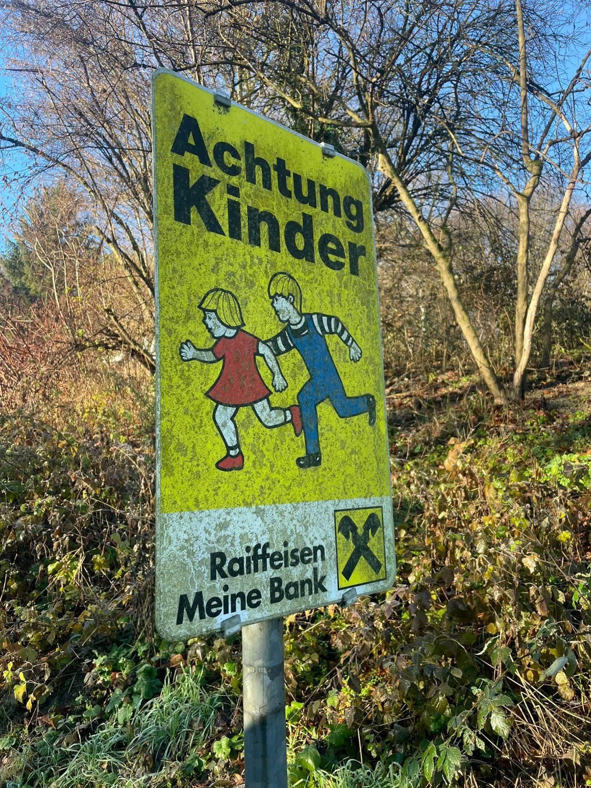 Ein gelbes Schild mit schwarzen Buchstaben sagt 'Achtung Kinder' und zeigt zwei laufende Kinder. Darunter steht 'Raiffeisen Meine Bank'. Der Hintergrund ist unscharf mit Bäumen und Pflanzen.