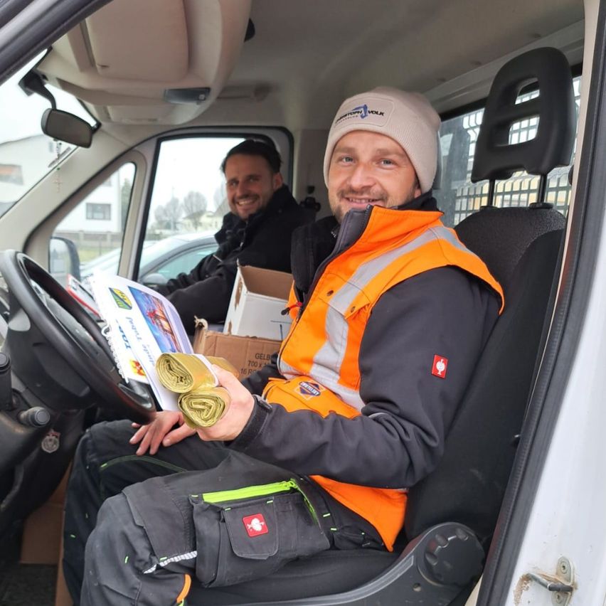 Zwei Männer sind in einem Fahrzeug, einer lächelt und hält ein gerolltes Papier, während der andere ein Buch betrachtet. Beide tragen Jacken mit reflektierenden Streifen.