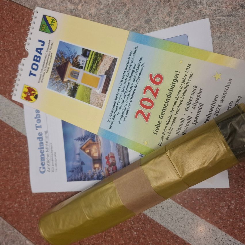 Eine goldumwickelte Papierrolle liegt auf einem weißen Papier und einem gelben Heft mit dem Text 'TOBAU' und 'Gemeinde Toba'. Das Heft hat ein Bild eines Hauses und eines Baumes auf seinem Cover.