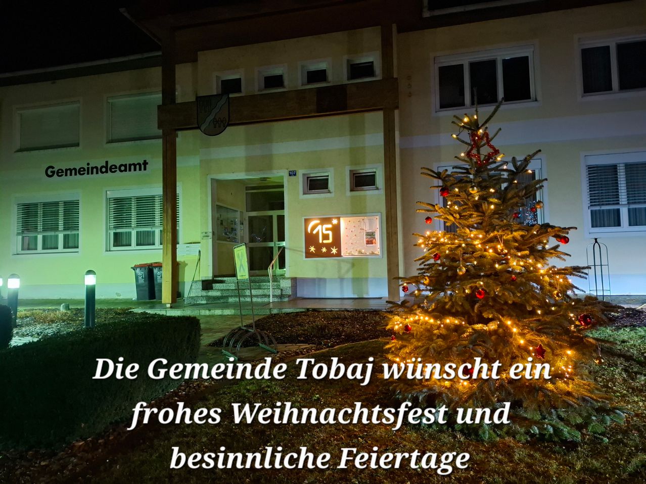 Ein Gebäude mit dem Text Eindeamt und einem beleuchteten Weihnachtsbaum mit Dekoration. Ein beleuchtetes Schild zeigt 15 Grad an.
