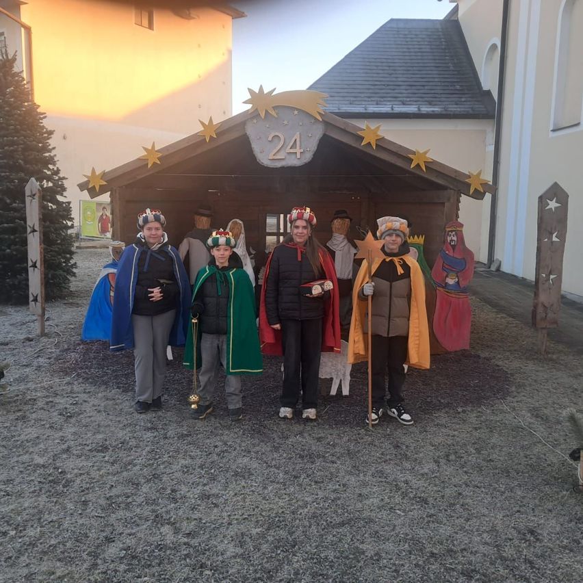 Fünf als Weise verkleidete Personen stehen vor einer Krippe mit der Nummer 24. Die Umgebung ist im Freien mit einem Gebäude, einem Weihnachtsbaum und Dekorationen im Hintergrund.