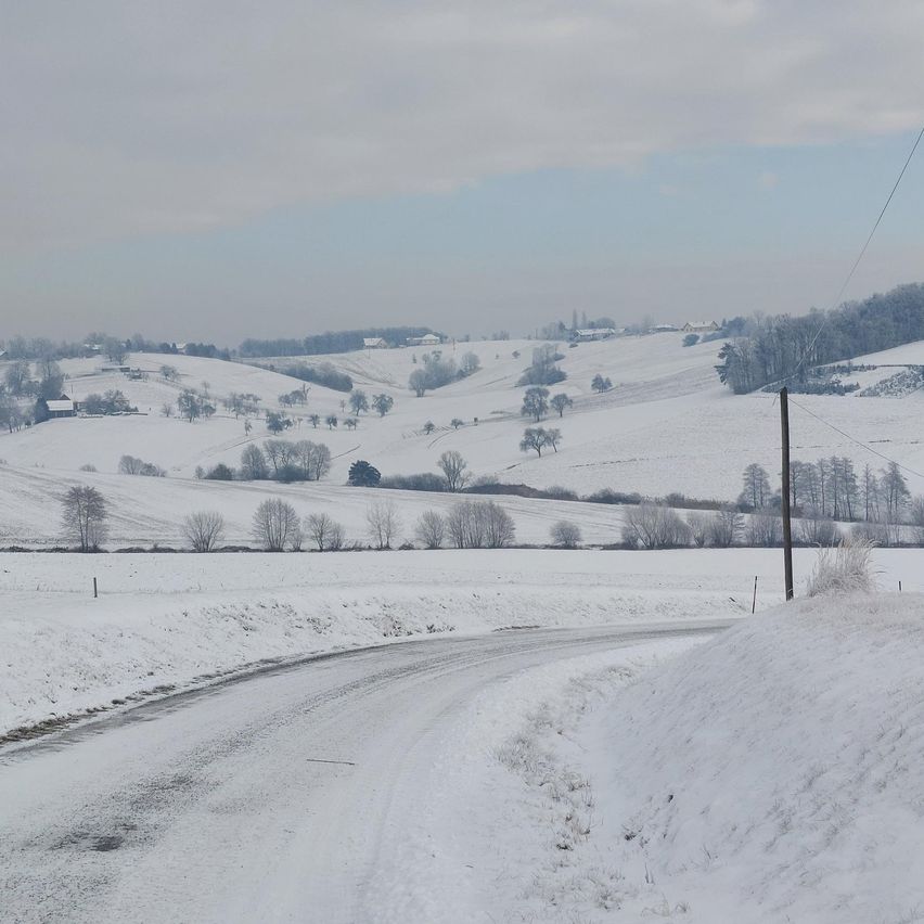 Eine verschneite Landschaft mit Hügeln und einer schneebedeckten Straße. Bäume und Häuser sind in der Ferne sichtbar. Der Himmel ist bewölkt.