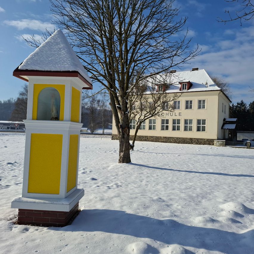 Eine gelbe und weiße Struktur steht auf einem verschneiten Feld, mit einem großen Gebäude, auf dem das Wort 'SCHULE' steht, hinter einem schneebedeckten Baum.