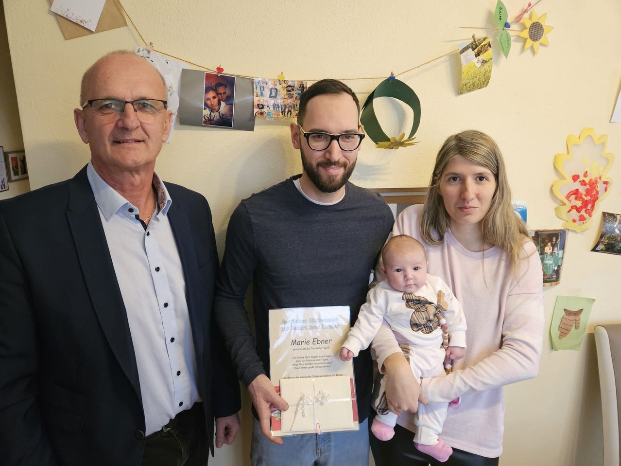 Drei Menschen stehen eng beieinander. Ein Mann hält ein Baby und eine Frau ein Zertifikat. Sie scheinen alle für ein Foto zu posieren. Der Raum ist mit einigen Papieren und Fotos dekoriert.
