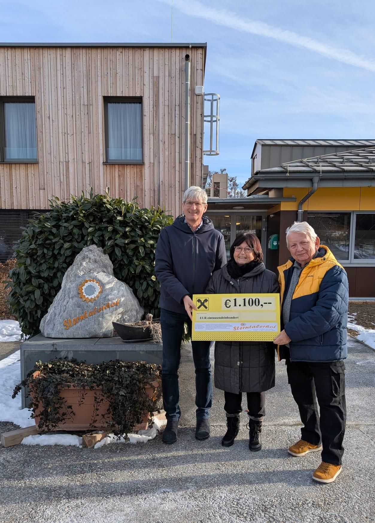 Drei Personen stehen vor einem Gebäude mit einem großen Felsen in der Nähe und halten ein gelbes Zertifikat über 1.100 Euro. Die Frau in der Mitte lächelt und schaut in die Kamera. Der Mann links trägt eine Brille und der Mann rechts trägt eine gelbe Jacke.
