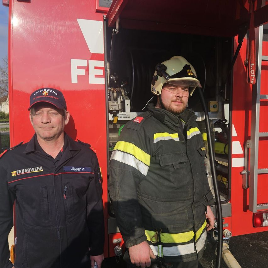 Zwei Feuerwehrleute stehen vor einem roten Feuerwehrwagen mit dem Wort FE darauf. Einer trägt eine Kappe und der andere einen Helm.