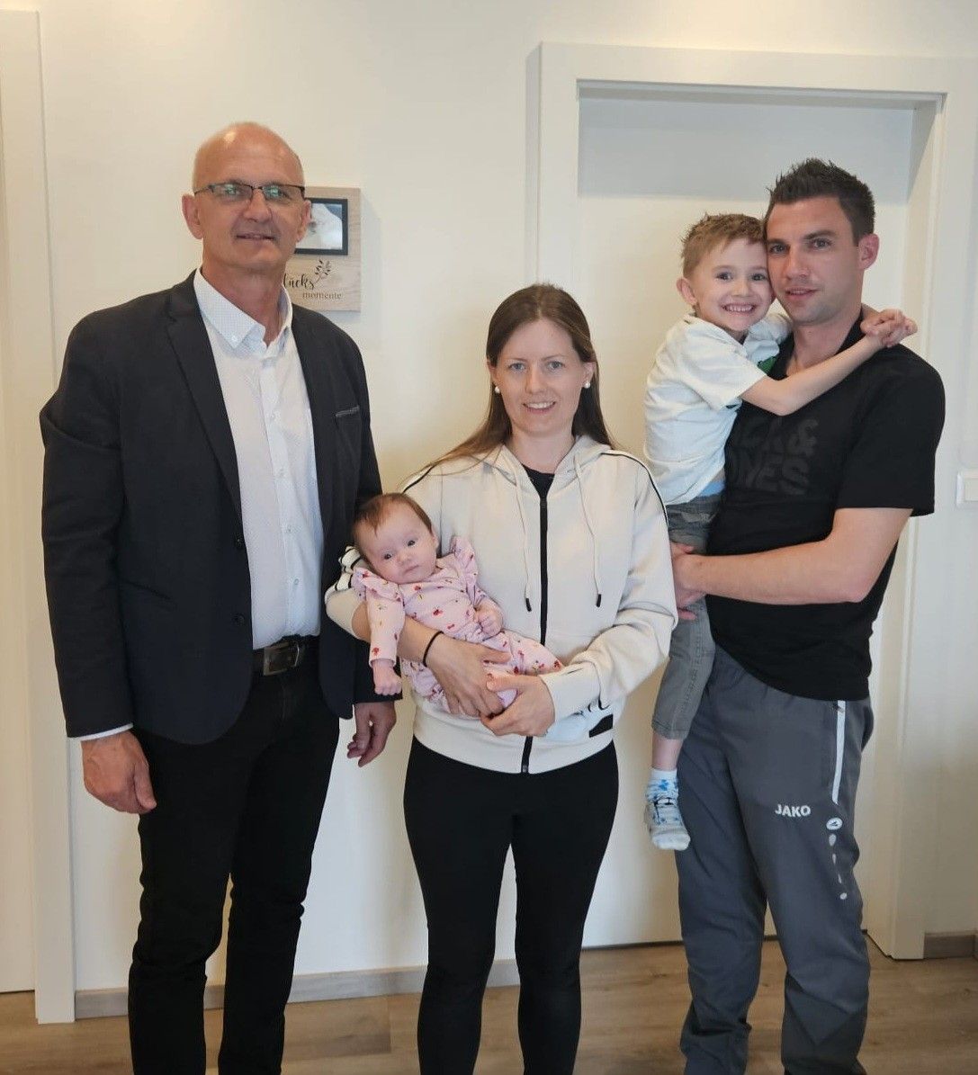 Eine vierköpfige Familie steht in einem Raum zusammen, mit einem Mann links, einer Frau, die ein Baby in der Mitte hält, und einem Mann rechts, der einen Jungen hält. Sie alle scheinen zu lächeln.