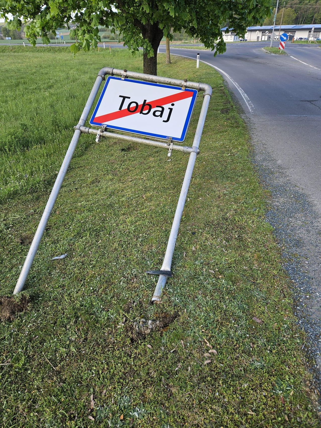 Ein beschädigtes Schild mit dem Wort 'Tobaj' liegt am Straßenrand auf dem Boden. Das Schild ist an einem Metallpfosten befestigt.