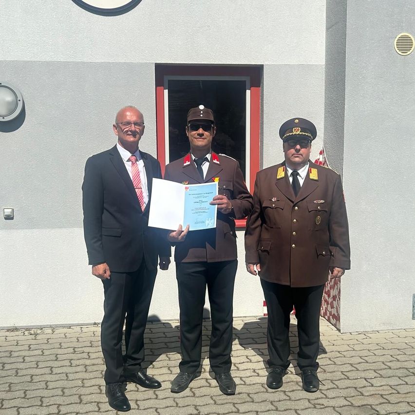 Drei Männer in Uniformen stehen vor einem Gebäude, einer hält eine Urkunde. Sie scheinen für ein Foto zu posieren.