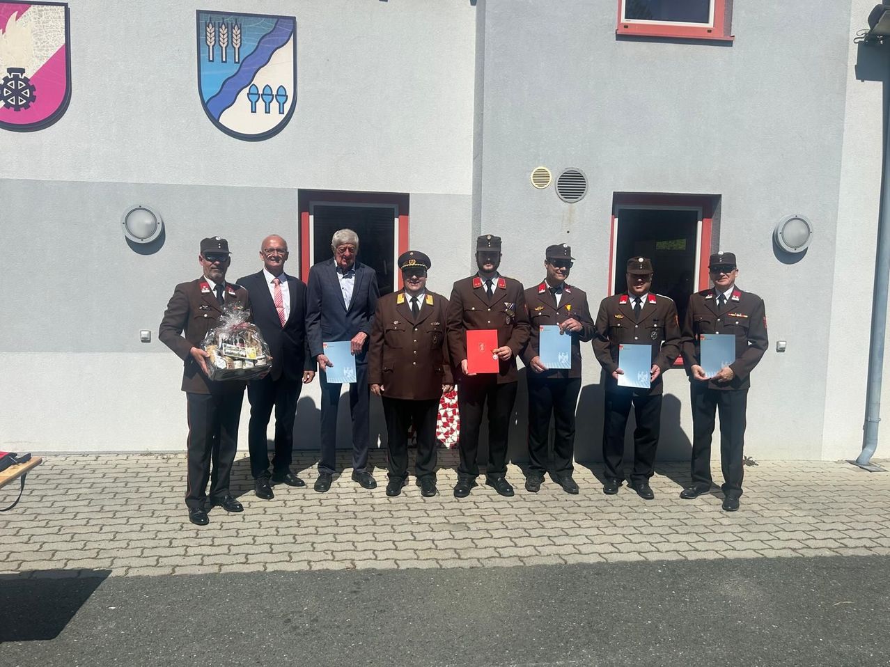 Eine Gruppe von Männern in Militäruniformen steht vor einem Gebäude. Ein Mann hält ein Geschenk, während andere Urkunden halten. Ein Logo ist an der Wand darüber angebracht.