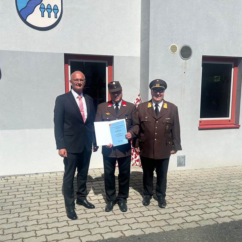 Drei Männer stehen vor einem Gebäude mit einem Zertifikat. Einer hält es. Sie tragen Uniformen und Anzüge. Hinter ihnen befindet sich ein Logo an der Wand.