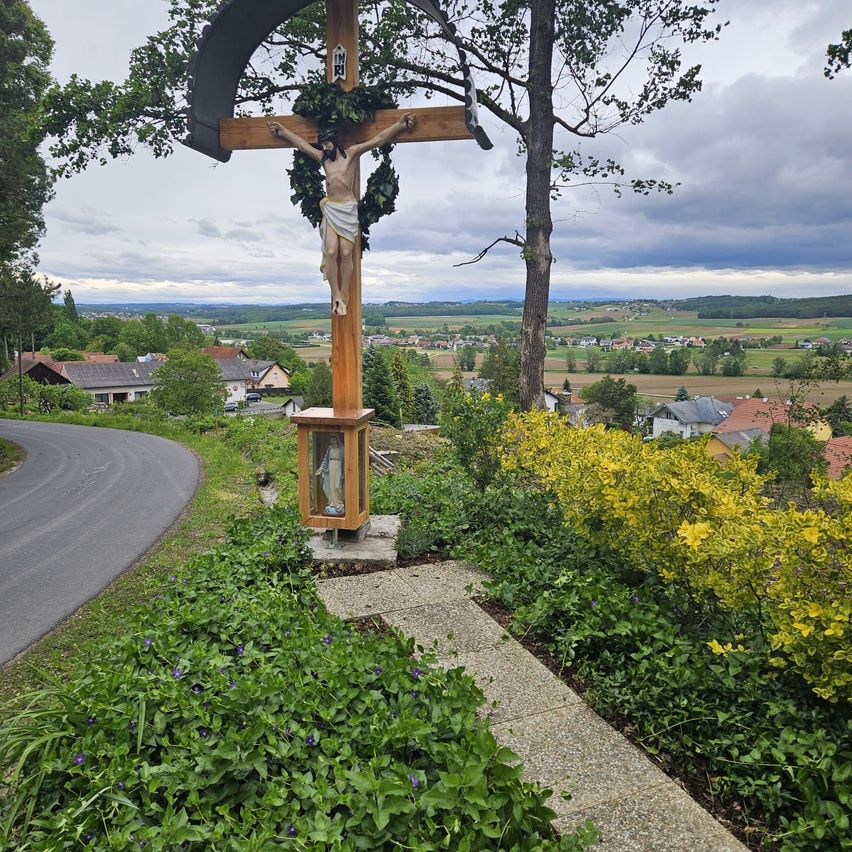 Ein Kruzifix steht auf einem Pfad mit Blick auf ein Dorf mit Häusern, Bäumen und einer Straße unter einem bewölkten Himmel.