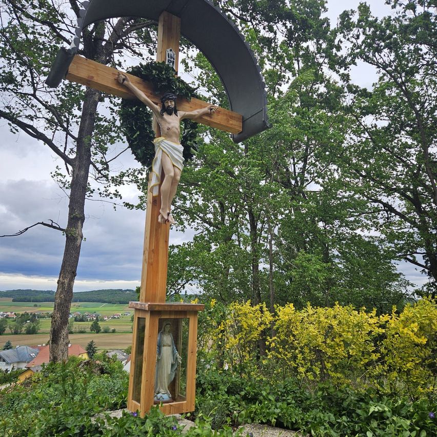 Ein Kruzifix steht in einem Garten mit Jesus am Kreuz und einer Statue der Maria darunter. Umgeben von Bäumen und Büschen bietet es einen Blick auf die Landschaft.
