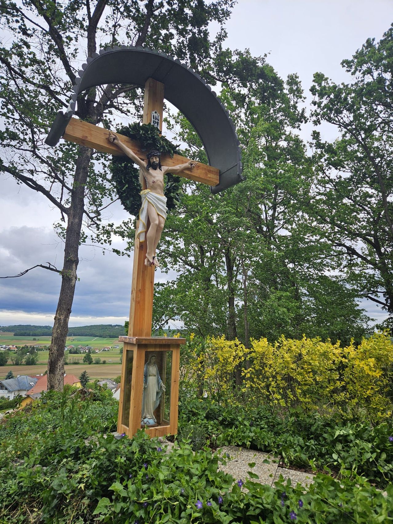 Ein großes Holzkreuz mit Jesus darauf steht in einem Garten. Dahinter sind Bäume und Büsche. Das Kreuz ist von grünem Laub umgeben.
