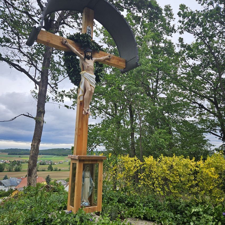 Ein großes Holzkreuz mit Jesus darauf steht in einem Garten. Dahinter sind Bäume und Büsche. Das Kreuz ist von grünem Laub umgeben.