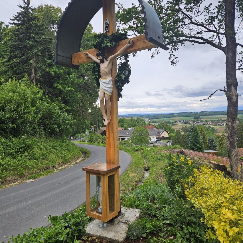 Ein hölzernes Kreuz mit einem Kruzifix, verziert mit grünen Blättern, steht am Straßenrand. Darunter befindet sich eine kleine Statue von Maria. Im Hintergrund ein kleines Städtchen mit Häusern und Bäumen.