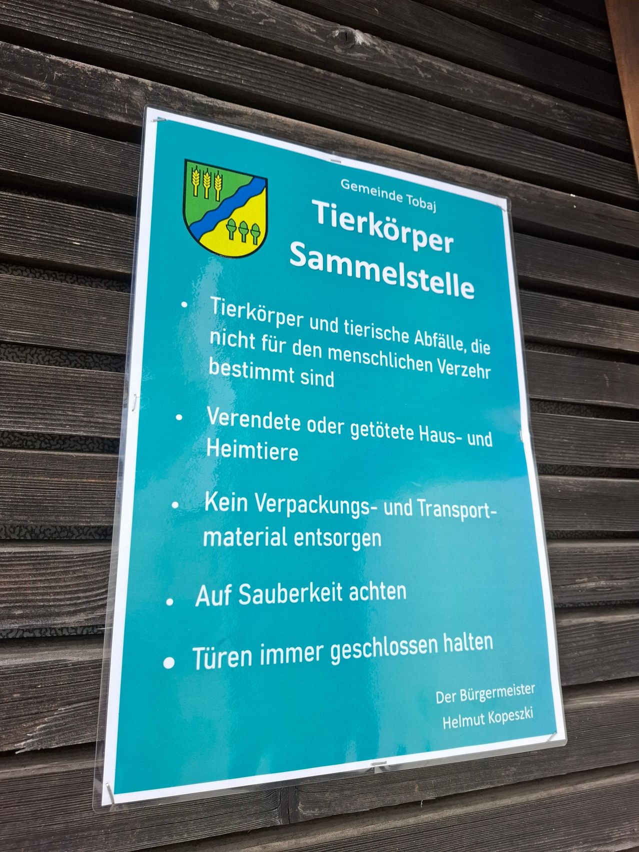 Ein Schild an einer Holzwand weist darauf hin, dass keine Tierkörper oder tierische Abfälle, die für den menschlichen Verzehr bestimmt sind, mitgebracht werden dürfen. Es warnt auch davor, Verpackungs- oder Transportmaterialien zu entfernen. Das Schild betont die Wichtigkeit der Sauberkeit und das geschlossen halten der Türen. Das Schild wurde vom Bürgermeister Helmut Kopezki herausgegeben.